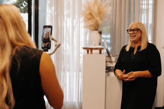 Two blonde women in a hair salon, one is filming on a phone with a gimbal and the other is smiling at the camera.