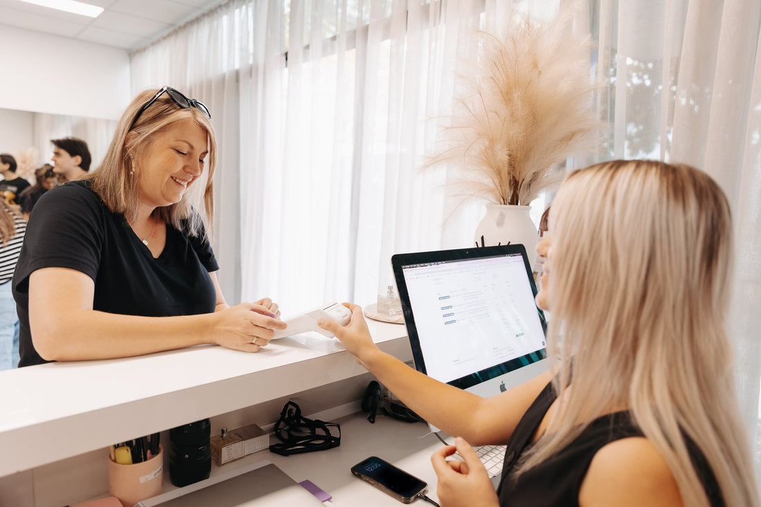 Two blonde women in a hairdressing salon, one behind the counter holding up an EFTPOS machine and the other, a client paying and smiling.