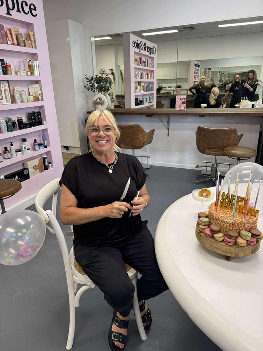 Owner of Ginger & Spice Hair Salon, Melinda Smith smiling at the camera while sitting in front of a cake with macarons, and balloons, celebrating the salon's 10th birthday.