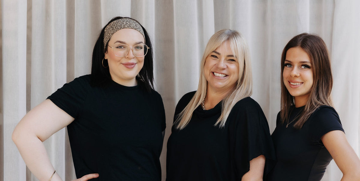 Three women standing side by side in matching black activewear or loungewear sets, posing with hands on hips in front of a light beige curtain backdrop.